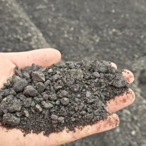 Handful of dark crushed rock or gravel held over a rough asphalt surface.