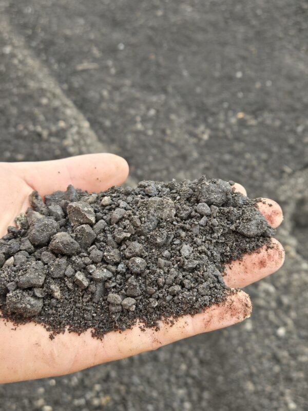 Handful of dark crushed rock or gravel held over a rough asphalt surface.