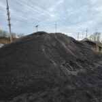 Large mound of black coal piled outdoors with power poles and lines in the background on a cloudy day.
