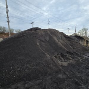 Large mound of black coal piled outdoors with power poles and lines in the background on a cloudy day.