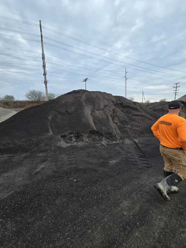 Worker in an orange shirt and tan pants stands near a large pile of coal on a gravel lot, with power lines and posts in the background.